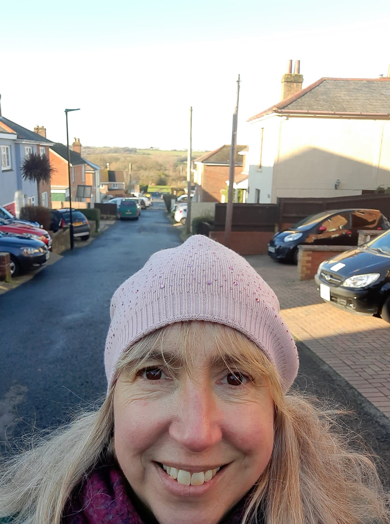 woman with long blonde hair wearing pink woolly hat with residential street behind her