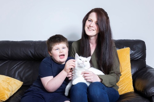 long-haired brunette woman with white cat on her lap sitting on black leather sofa with yellow cushions while young short-haired brunette boy sits next to her