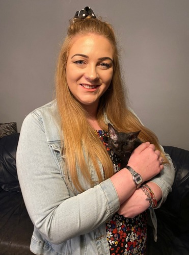 long-haired blonde woman wearing denim shirt and holding black one-eyed Cornish Rex kitten