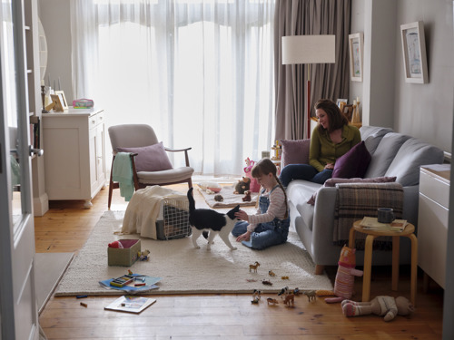 an adult white woman with brunette hair sitting on a grey sofa while a young girl with brunette hair sitting on the floor stokes a black-and-white cat