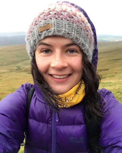 selfie of a brunette woman wearing a knitted grey beanie hat, purple coat and yellow Cats Protection scarf while outdoors in front of greenery